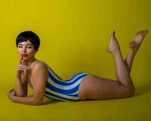 Wall Mural - Attractive brunette lies on her stomach in a striped bikini. Short-haired young woman in bodysuit on a yellow background in the studio.