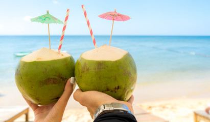 Two fresh green coconuts for drinking in male and female hands with straw against the backdrop of a paradise tropical beach.Wedding,honeymoon, vacation and travel to tropical countries or Womens Day.