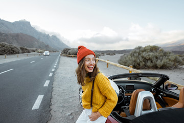 Portrait of a joyful woman dressed in bright sweater and hat enjoying road trip, standing near the car on the roadside of the picturesque volcanic valley on a sunset