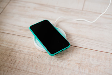 Smartphone in mint silicone case is charged from a wireless charger. The mobile phone is charged on a wooden nightstand or shelf.