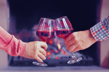 Man and woman hand toasting wine glasses in front of lit fireplace at home. Elderly couple have romantic dinner with red wine over fireplace background. Romantic winter night for couple in love.