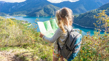 An Independent Blonde Girl With A Tourist Backpack Lays A Route On A Map On The Shore Of A Mountain Lake In The Italian Alps. Picturesque Landscape On A Blue Mountain Lake.
