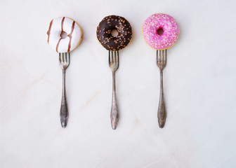 Glazed, chocolate frosted and sprinkled donuts on forks isolated on marble background. Top view. Forks Madness set