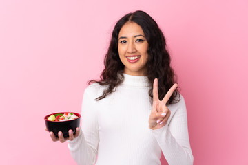 Wall Mural - Mixed race woman holding a bowl full of noodles over isolated pink background smiling and showing victory sign