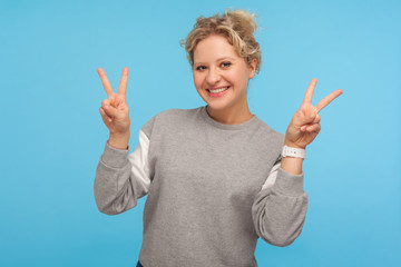 Excited cheerful woman with short curly hair in sweatshirt showing victory gesture, double peace sign, rejoicing at successful results, goal achievement. indoor studio shot isolated on blue background