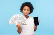 © khosrork - Adorable cute surprised little boy with curly hair in T-shirt holding lots of money and cell phone, looking at camera with surprised shocked expression. indoor studio shot isolated on blue background