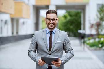 Attractive smiling caucasian elegant businessman in suit and with eyeglasses standing outdoors, holding tablet and looking at camera.