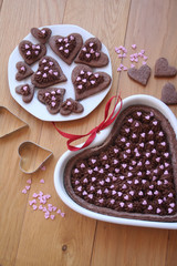 Wall Mural - Dark chocolate tart in shape of a heart with a plate of decorated cookies on wooden table with decorations. Valentine's day background