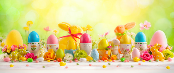 Wall Mural - Easter concept with colorful decorated eggs in egg cups, rabbits and flowers on white wooden table on spring nature background.