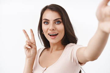 Close-up portrait friendly beautiful caucasian woman taking selfie in kawaii pose, holding camera with extended arm and make peace sign, smiling upbeat, live stream from concert, white background