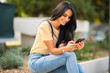 © mimagephotos - smiling young woman sitting outside looking at phone