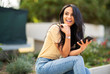 © mimagephotos - young woman sitting outside holding cellphone and smiling