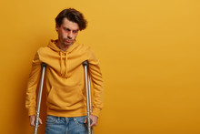 Frustrated Man With Injured Face Learns To Walk Again, Needs To See Surgeon, Stands On Crutches, Has Recovery Period After Terrible Accident, Concentrated Down, Isolated On Yellow Wall, Blank Space