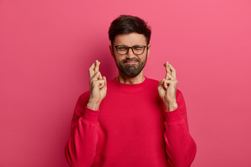 Wall Mural - Bearded man crosses fingers, hopes for best, presses lips, believes in good luck, wears spectacles and red sweater, stands against pink wall, has big desire, dreams to achieve goal, aspires something