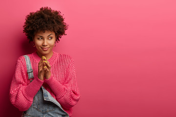 Cunning young Afro American woman steepls fingers, looks aside, has intention or tricky plan, wears knitted sweater and denim overalls, isolated on pink background, prepares surprise for husband