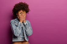 Isolated Shot Of Shocked Frightened Curly Woman Peeks Through Fingers, Stares With Widely Opened Eyes, Wears Denim Jacket, Hides Face From Someone, Isolated Over Purple Wall, Copy Space Aside