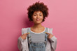 Boastful curly Afro American woman got good job, points at herself, talks about personal achievement, becomes champion, wears denim dungarees and anorak, isolated over rosy pastel background