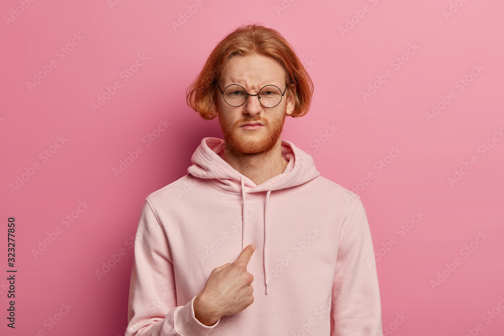 Displeased red haired man smirks face and points at himself, feels bothered and unhappy, asks why me, being accused in doing something wrong, dressed in hoodie, isolated over rosy pastel wall - obrazy, fototapety, plakaty 