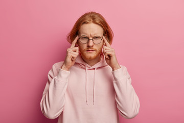 Wall Mural - Portrait of intense redhead man presses index fingers on temples, focused on task, tries to concentrate on difficult task, has ginger thick beard, wears hoodie, isolated over pink background
