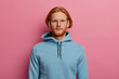 © WHstudio Leushin N - Serious looking confident bearded man with ginger hair, looks straightly at camera with calm expression, wears blue hoodie, isolated over rosy pastel background. Human face expressions concept