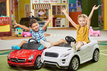 Wall Mural - Happy kids sitting in toy cars in nursery school. Cute little boy and girl giving five each other in playroom. Fun in kids center.