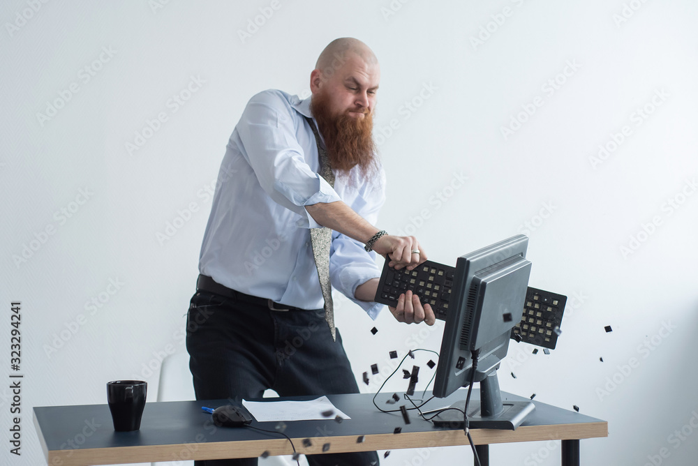 Angry bald man with a red beard in the office in a business suit crashes a computer. Manager with a nervous breakdown breaks the keyboard on the monitor. - obrazy, fototapety, plakaty 