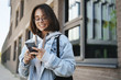 © Liubov Levytska - Young queer girl, college student standing on street, order taxi using application on smartphone, smiling as texting friend, meeting with team outside campus, scrolling news feed in social media