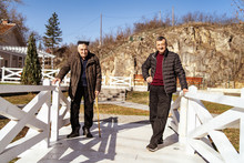 Two Men Senior Man Father And Son 80 And 60 Years Old Standing On White Bridge In Sunny Winter Or Spring Day Posing Looking To The Camera By The House In Village