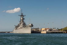Oahu, Hawaii, USA. - January 10, 2012: Pearl Harbor. Bow Of Gray USS Missouri Docked And Used As Historic Museum On Blue Water And Under Blue Cloudscape. Brown Buildings On Shore.