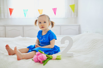 Wall Mural - Adorable toddler girl in blue dress sitting on bed with pink hyacinths and white wooden number two