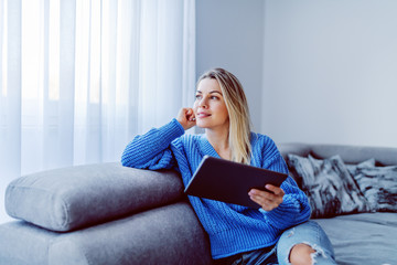 Beautiful caucasian blond young woman sitting on sofa in living room, holding tablet and looking trough window.