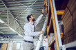 Full length of Caucasian bearded smiling graphic engineer standing on ladder and trying to reach box. Printing shop interior.
