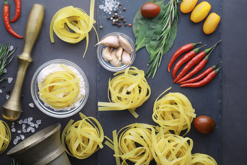 Wall Mural - Pasta on the table with spices and vegetables. Noodles with vegetables for cooking on a black stone background.