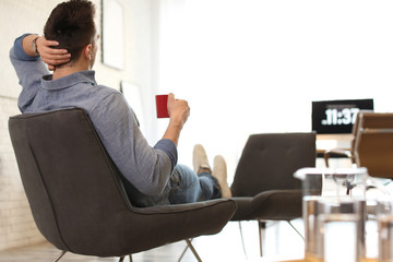 Wall Mural - Young man with cup of drink relaxing in comfortable chair at workplace