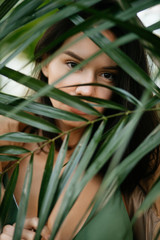 Wall Mural - young caucasian woman with charming look, lady with black beautiful eyes look at camera, cover the face behind green exotic plant, green leaves of tree. mysterious woman in rainforest