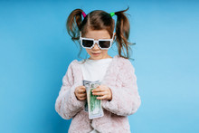 Portrait Of Funny, Cool, Glamorous Little Caucasian Kid Girl In Sunglasses Isolated Over Blue Background. Cool Child In Pink Coat, With Pigtails On Head Stand Holding Dollar Bills In Hands.