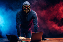 Portrait Of An Anonymous Man Hiding His Face Behind A Scary Neon Mask In A Colored Smoke. Male In Pullover Stand In Scary Mask Isolated Over Dark Smoky Space. Modern Devices, Laptop On Table.
