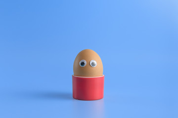 One brown egg with eyes in a round red box on a blue background. Minimal Happy Easter concept decoration. Copy space for text mockup.