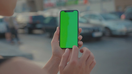 Lviv, Ukraine - May 19, 2018: Close-up of young urban girl showing vertical smartphone with greenscreen chrome key technology on parking lot outside. City life and modern gadgets.