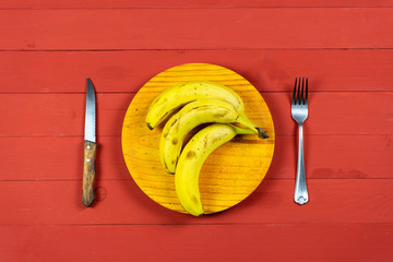 Ripe bananas on a wooden plate with cutlery on a red wooden table