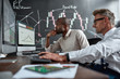 © Svitlana - Planning for secure future. Two diverse colleagues traders discussing ideas while sitting in the office in front of multiple computer screens. Blackboard full of data analyses in the background.