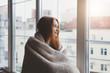 © zakalinka - beautiful thoughtful pensive calm smiling young woman looking through the window at home wrapped in warm comfy blanket