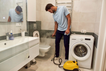 Wall Mural - Professional at work. Young caucasian man in uniform cleaning toilet with steam cleaner. Male cleaner working in the bathroom