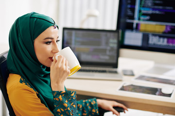 Wall Mural - beautiful young muslim woman in hijab sitting at office table and drinking mug of coffee