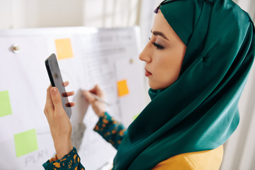 Wall Mural - beautiful muslim project manager checking data on smartphone screen when writing on whiteboard and p