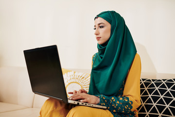 Wall Mural - muslim female freelance programmer sitting on sofa at home and working on laptop