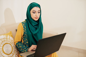 Wall Mural - beautiful muslim young woman in traditional dress and hijab sitting on sofa and working on laptop at