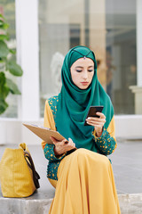 Wall Mural - serious young muslim businesswoman in traditional dress sitting outdoors and checking text messages 