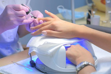 Manicurist Master Is Covering Painting Client's Nails Top Coat Shellac, Hands Closeup. One Hand Drying In UV Lamp. Professional Manicure With Gel Polish In Beauty Salon. Beauty Industry Concept.