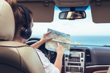 Woman driver in the headphones driving a car and looking at th map. Girl relaxing in auto trip traveling along ocean tropical beach in background. Traveler concept.